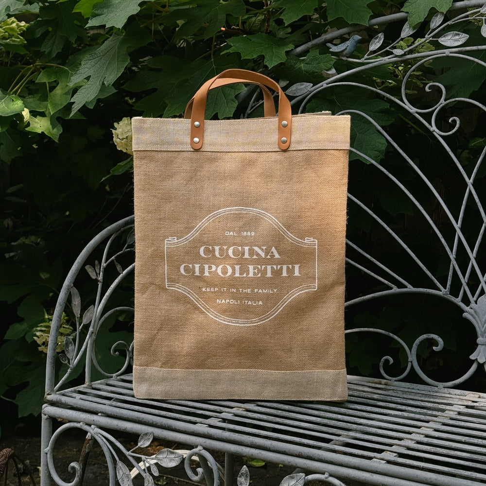 Front view of Cucina Cipoletti market tote showing printed logo, leather handles and jute canvas on an iron garden bench