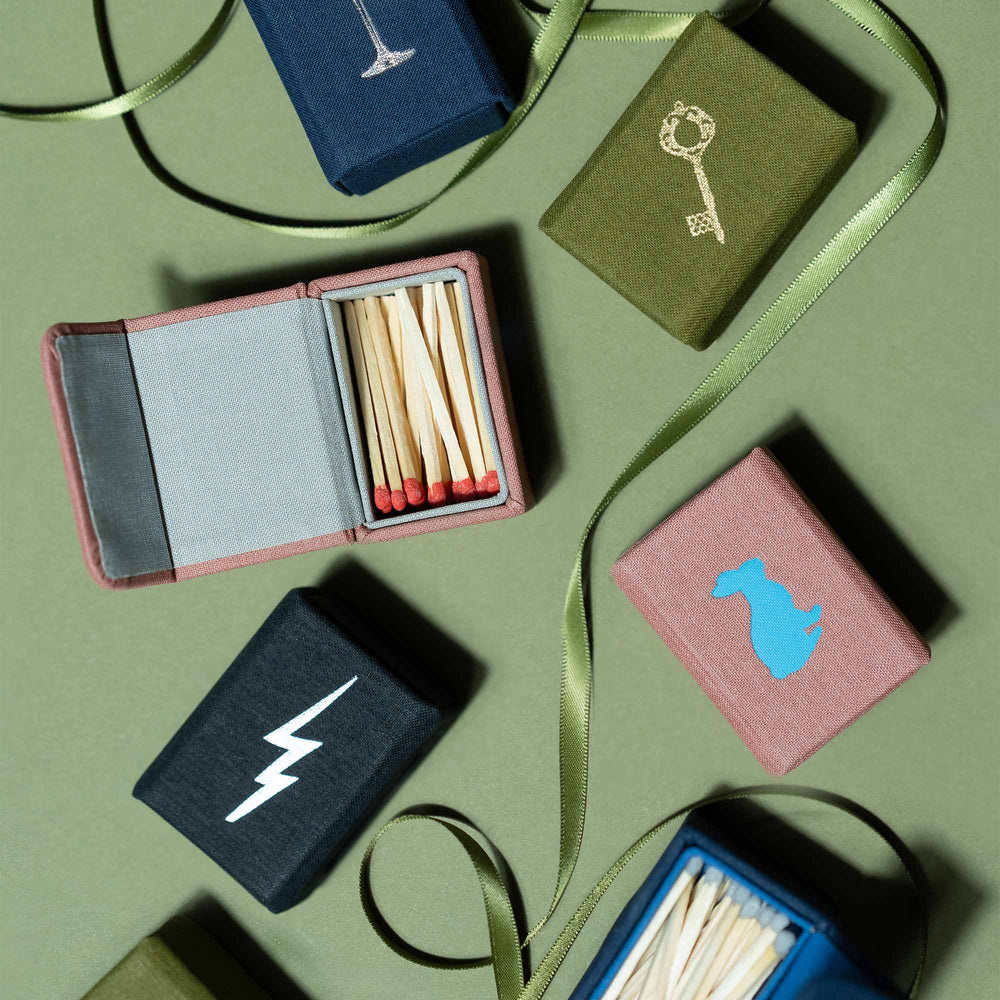 Top-down view of Toast matchbox open with red-tipped matches beside closed fabric-covered matchboxes on green background
