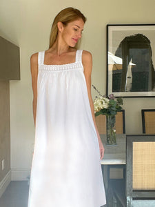 Woman wearing a white sleeveless dress in a room with a painting and vase in the background.