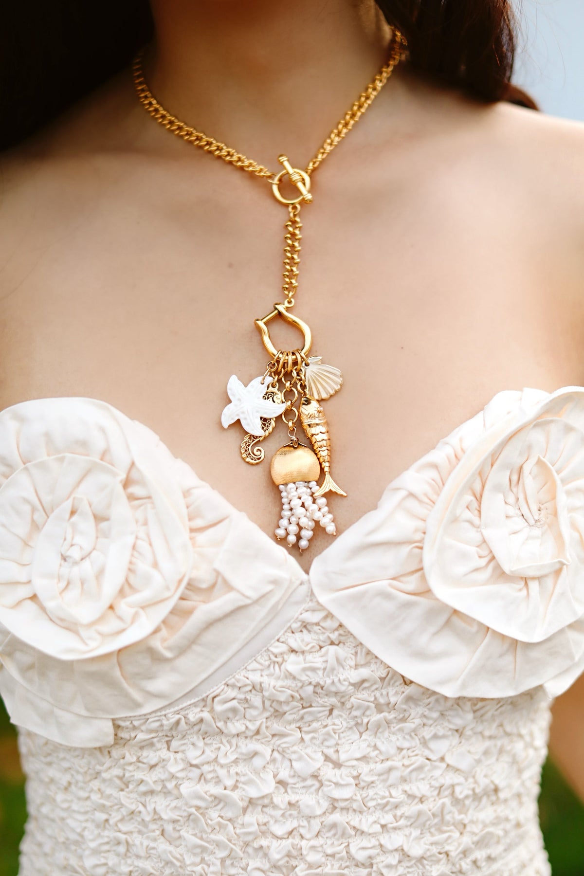 Close-up of Mermaid’s Memento necklace with gold chain, toggle clasp and sea charms with pearl tassel on a cream ruched top