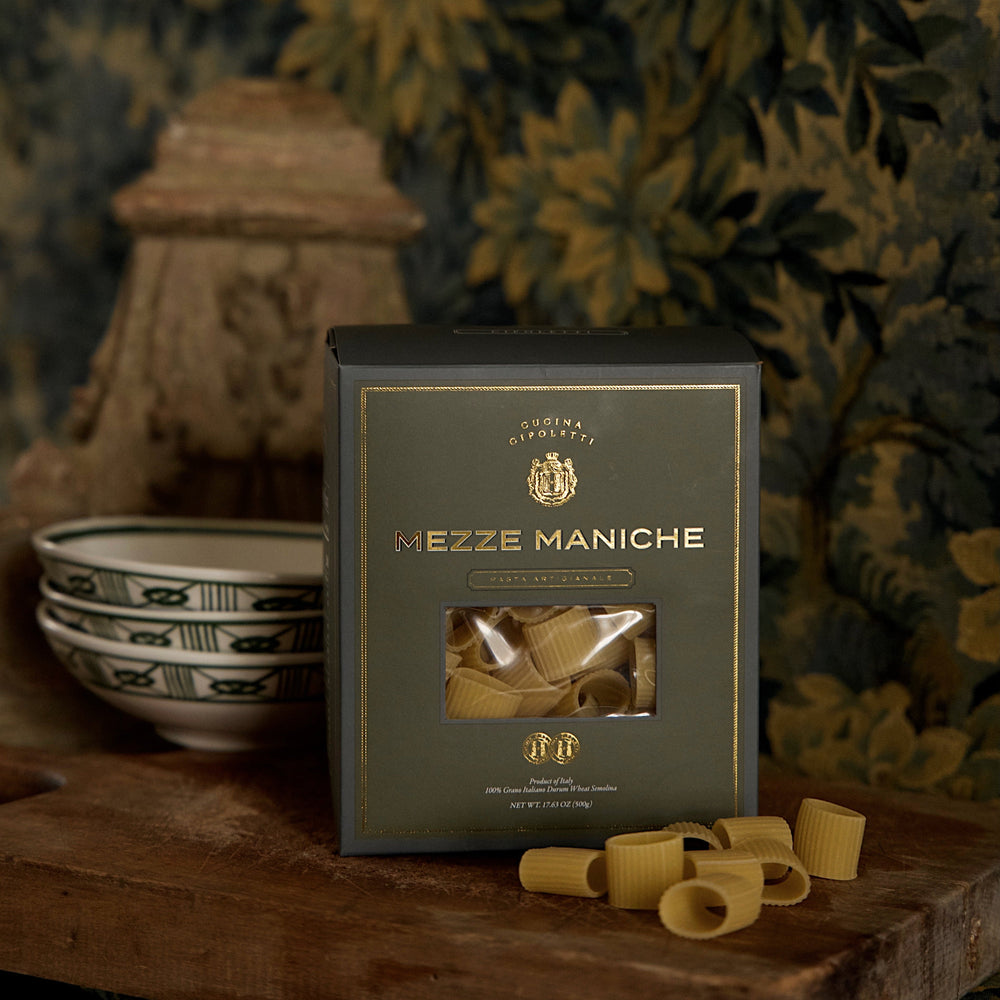 Front view of Mezze Maniche pasta box showing gold-accented green packaging with clear window revealing rigatoni on table