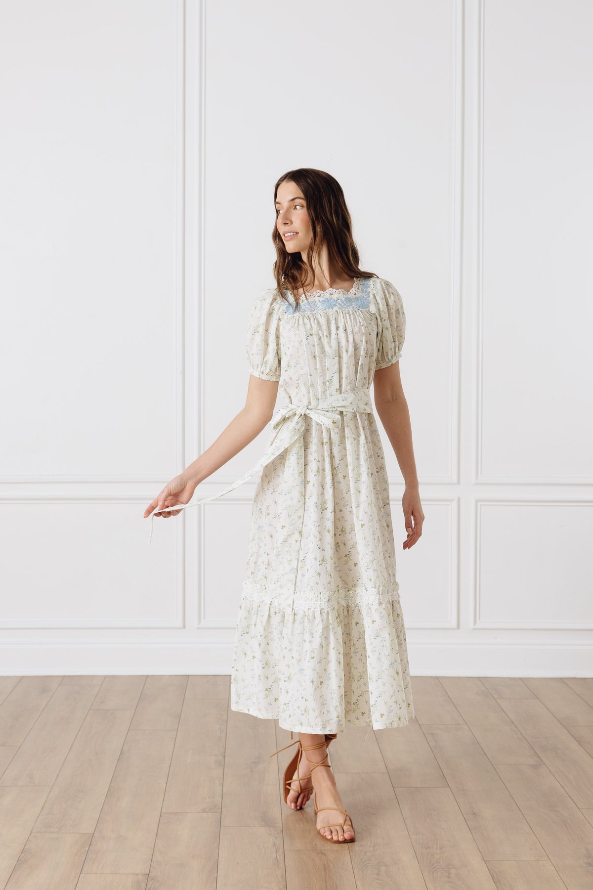 Three-quarter view of OTM Exclusive Maria Dress showcasing floral embroidery, puff sleeves, and removable sash on a model against a white paneled wall