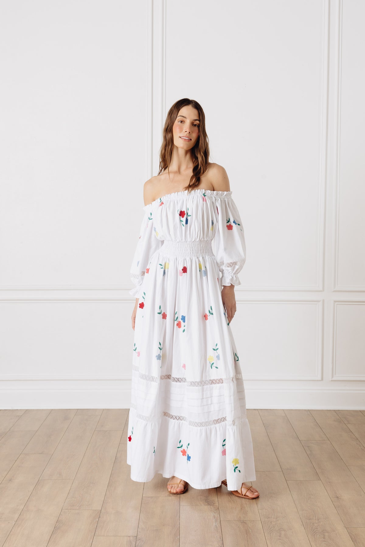 Front view of Rosalia Long Dress showcasing off shoulder styling, floral embroidery, and lace trim against a white wall