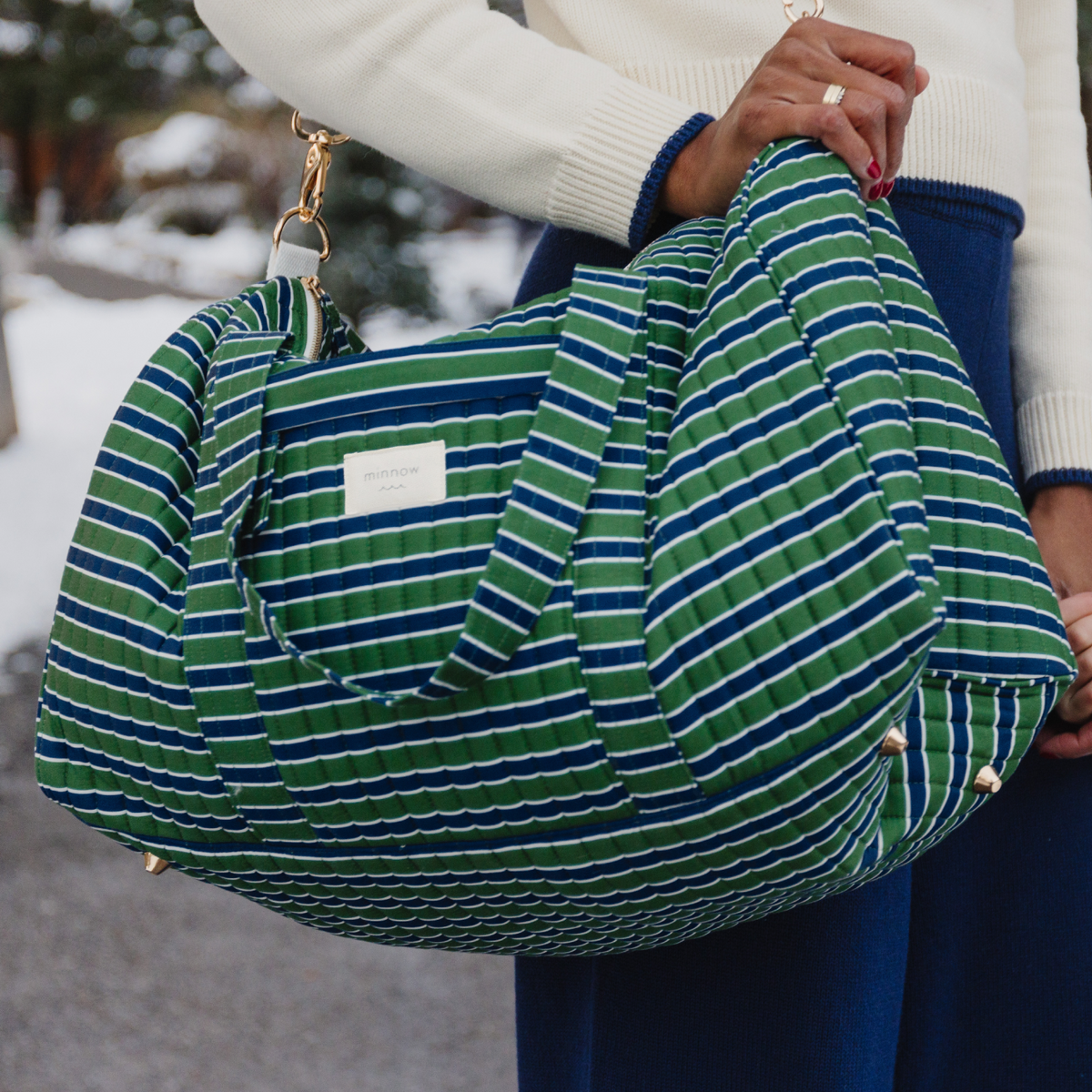 Angled view of Evergreen Stripe Weekender Bag showcasing green and navy stripes and gold hardware with snowy background