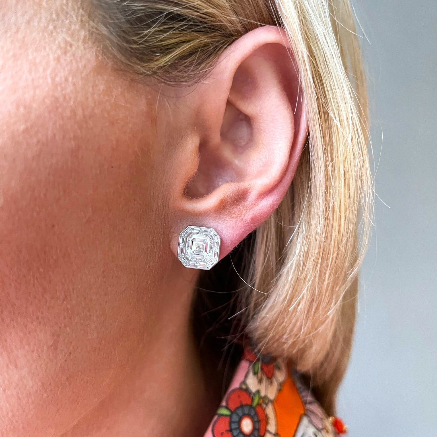 Close-up of a woman's ear wearing Mosaic Studs with Asscher Cut Diamonds in white gold. She has blonde hair and a colorful floral top. The background is softly blurred.