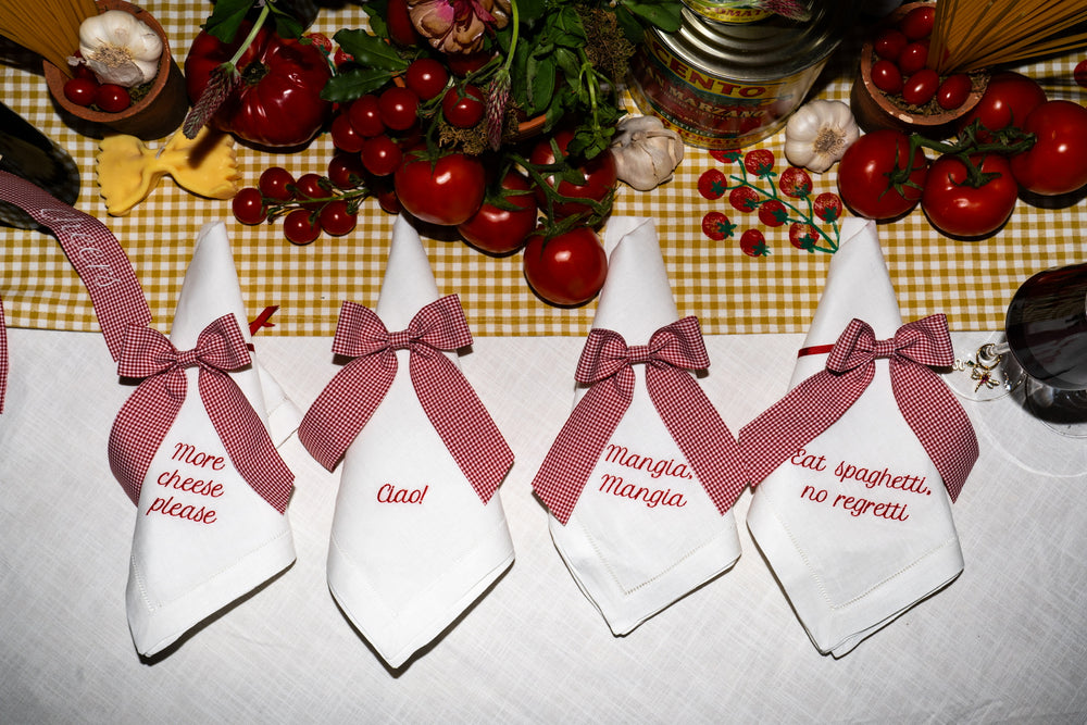 Top view of Gingham Napkin Bow showcasing red gingham pattern tied around white cloth napkins with a yellow gingham runner and tomatoes in the background