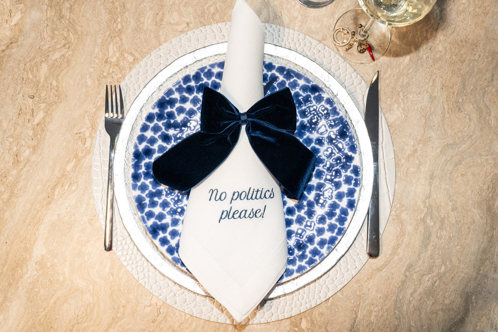 Top-down view of Mixed Messages dinner napkin showcasing embroidered no politics please message and navy velvet bow on a blue patterned plate