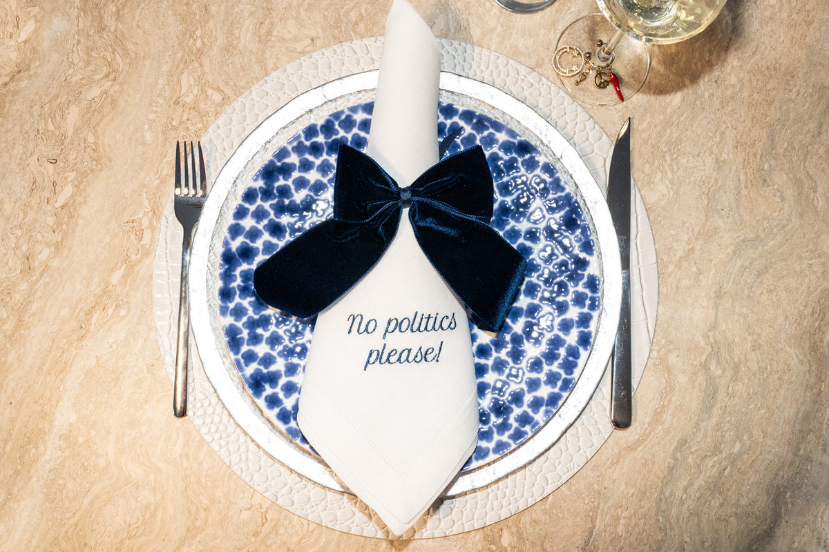 Top-down view of Mixed Messages dinner napkin showcasing embroidered no politics please message and navy velvet bow on a blue patterned plate