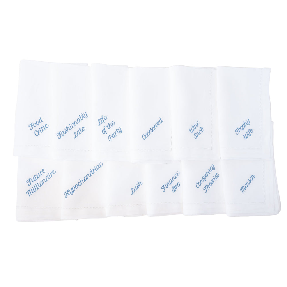 Top view of Party Archetype dinner napkins showcasing blue embroidered personality titles on white linen background