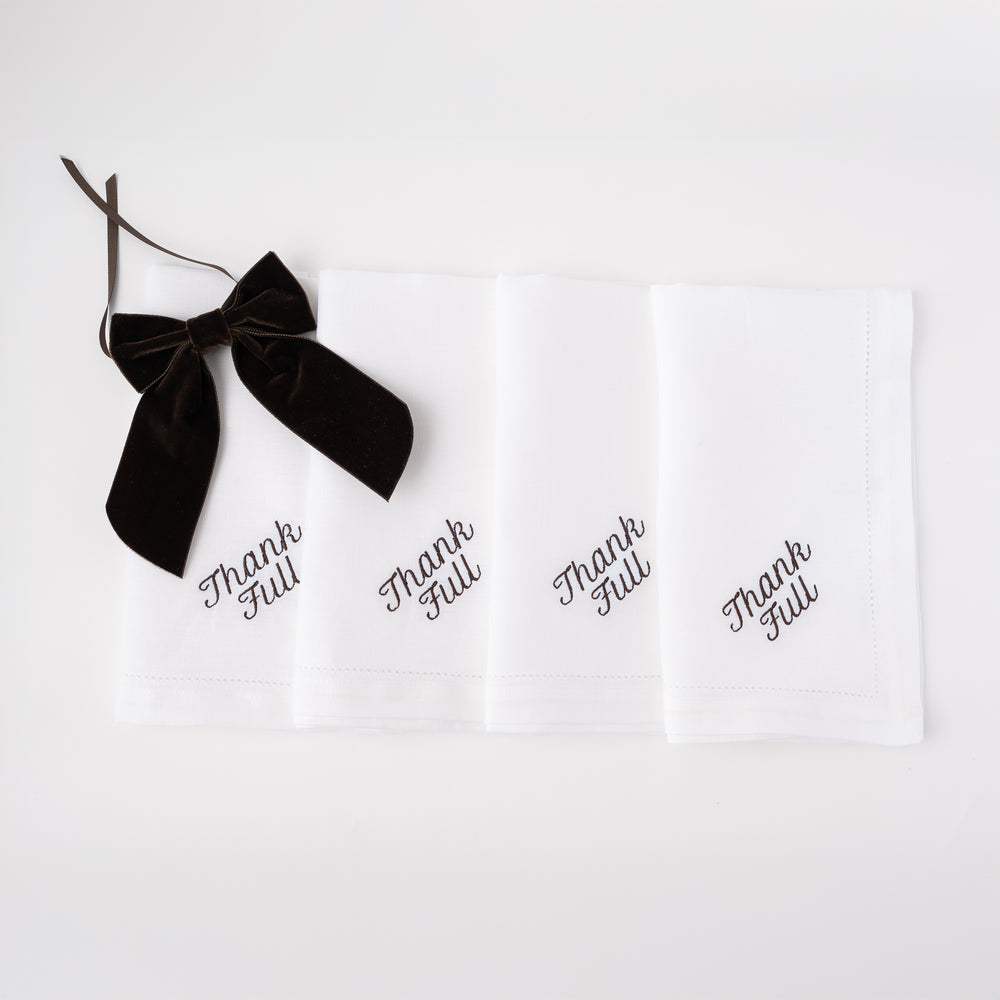 Front view of white linen dinner napkins showcasing black embroidered Thank Full script and a black velvet bow on white background