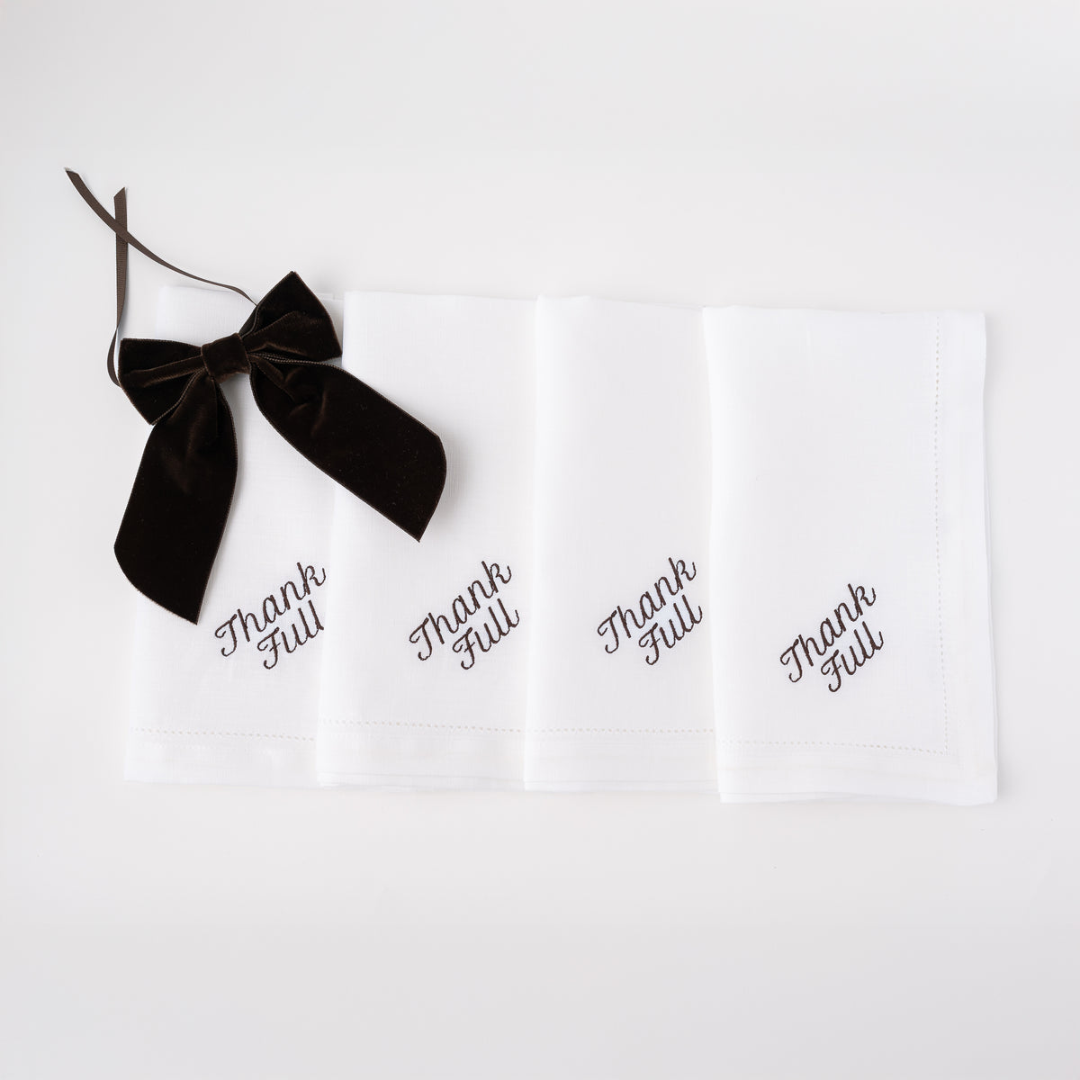 Front view of white linen dinner napkins showcasing black embroidered Thank Full script and a black velvet bow on white background
