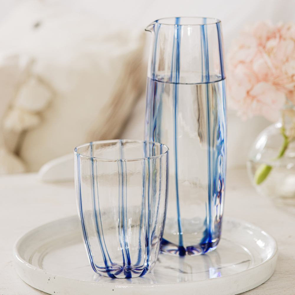 Front view of Nuovo Stripe blue glass pitcher with matching tumbler, showing vertical painterly blue stripes on a white tray