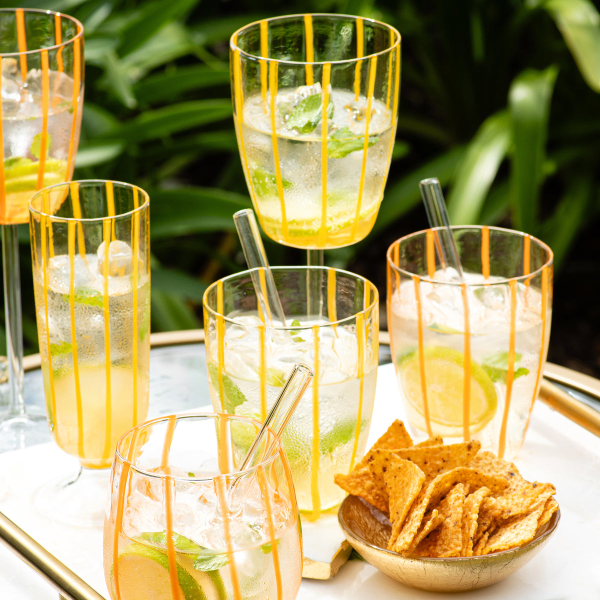 Angled view of Nuovo Stripe wine glass showing hand painted yellow vertical stripes on a tray with iced drinks and chips