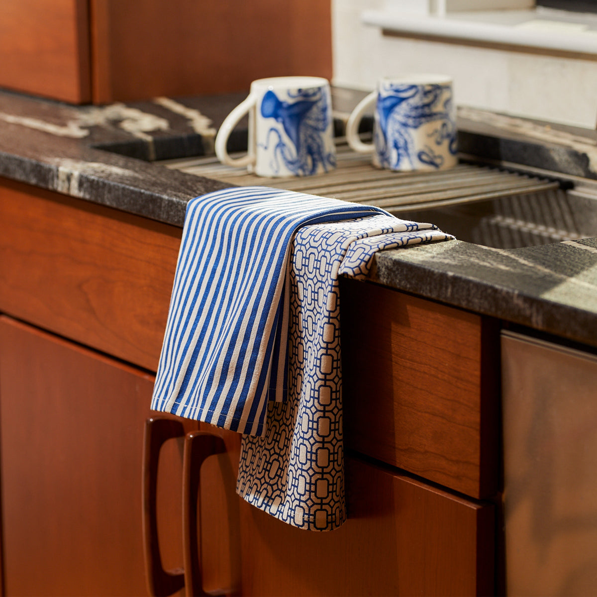 Two blue cotton kitchen towels draped over a sink edge, one pinstripe and one geometric, with two blue-and-white mugs on the countertop