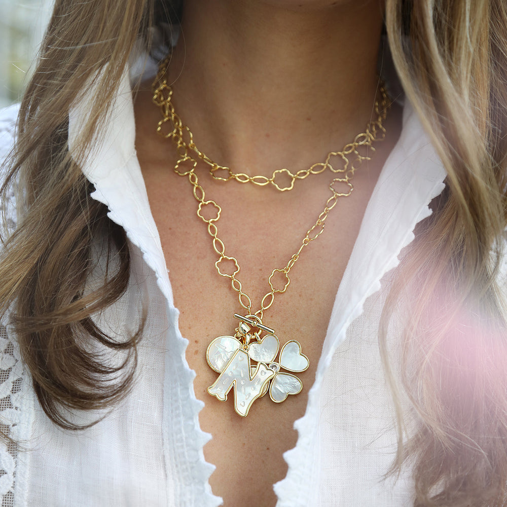 Close-up of Letter Charm pendant with mother-of-pearl letter N and quatrefoil charms on layered gold chain against white blouse