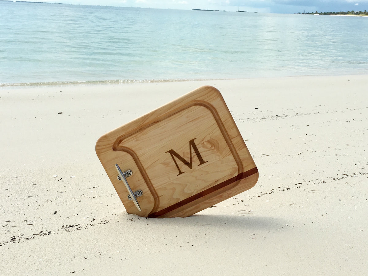 Angled view of Nautical Cleat Appetizer Board showing chrome cleat handle, recessed juice groove, mahogany inlay and engraved M on a sandy beach