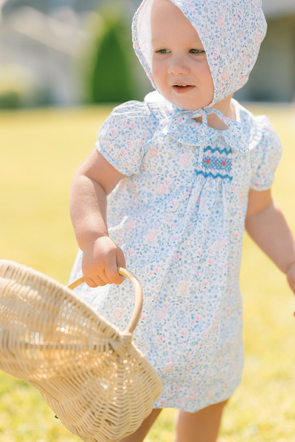 Three-quarter front view of Olympe Baby Set in floral blue on a toddler showing smocked front, puff sleeves, and matching bonnet in a sunlit yard