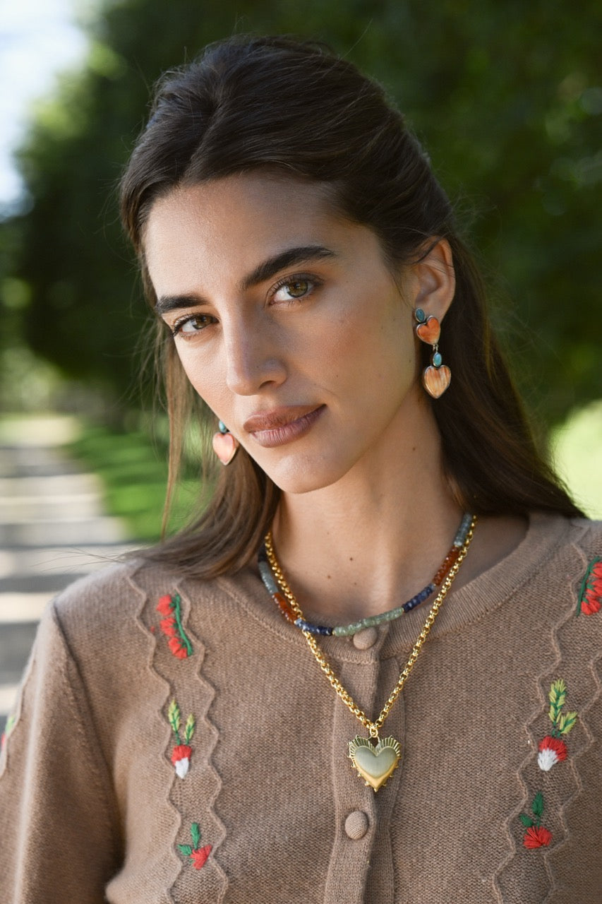 Close-up of camel Chelsea cardigan showing floral embroidery and scalloped trim, blurred greenery background