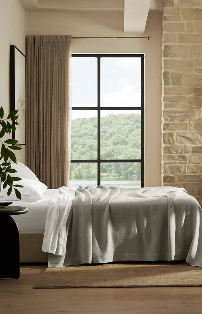 Bed with pewter Essential Bed Blanket casually draped over the edge in a sunlit bedroom with large window and stone wall