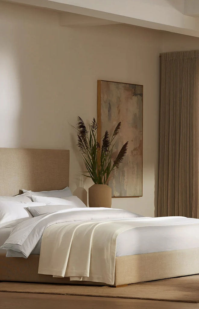 Bed with cream Essential Bed Blanket draped at the foot in a neutral bedroom with beige headboard and vase of dried grasses
