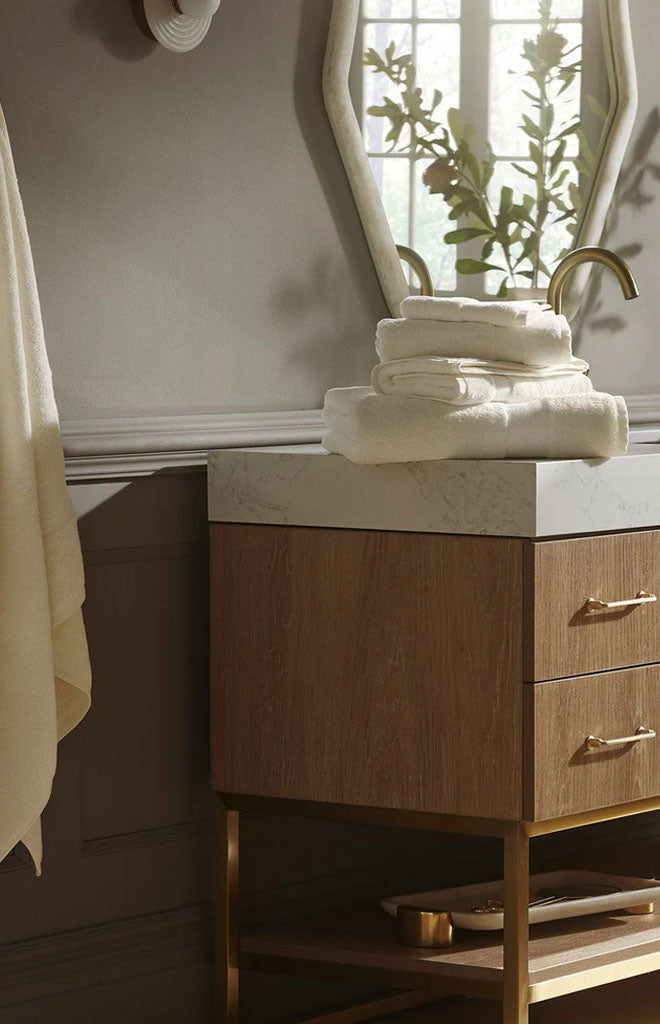Stack of cream plush bath sheets folded on a marble-top wooden vanity with brass faucet and hexagonal mirror