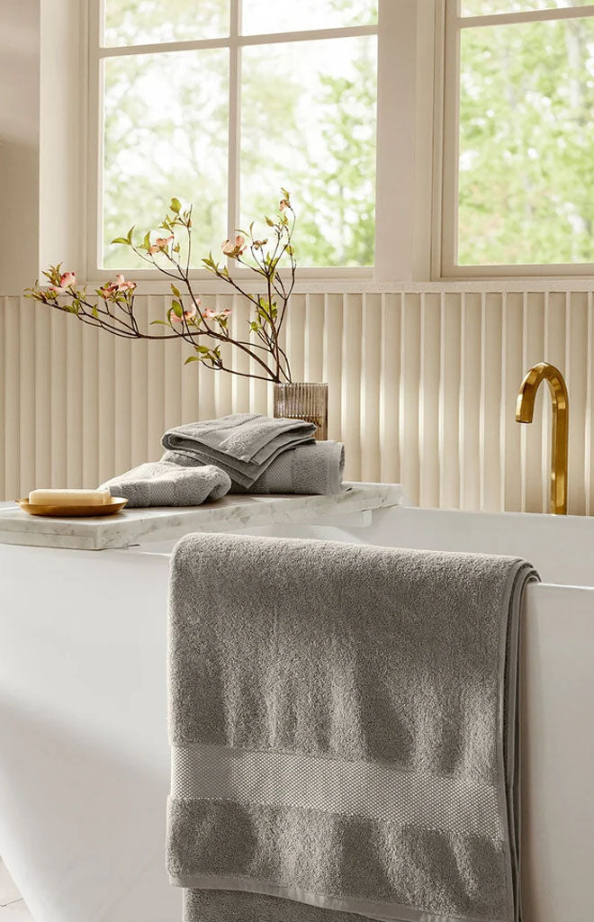 Pewter bath towels draped over a freestanding bathtub with folded towels on a marble tray and a gold faucet