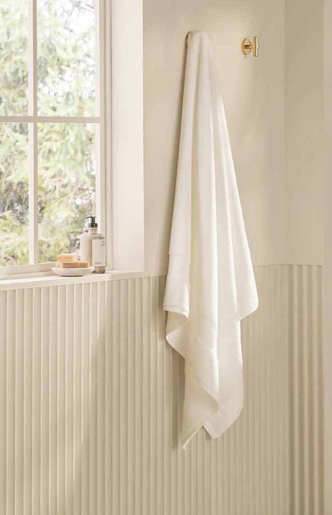 White plush bath sheet hanging from a brass hook in a bright bathroom next to a window with soap and lotion
