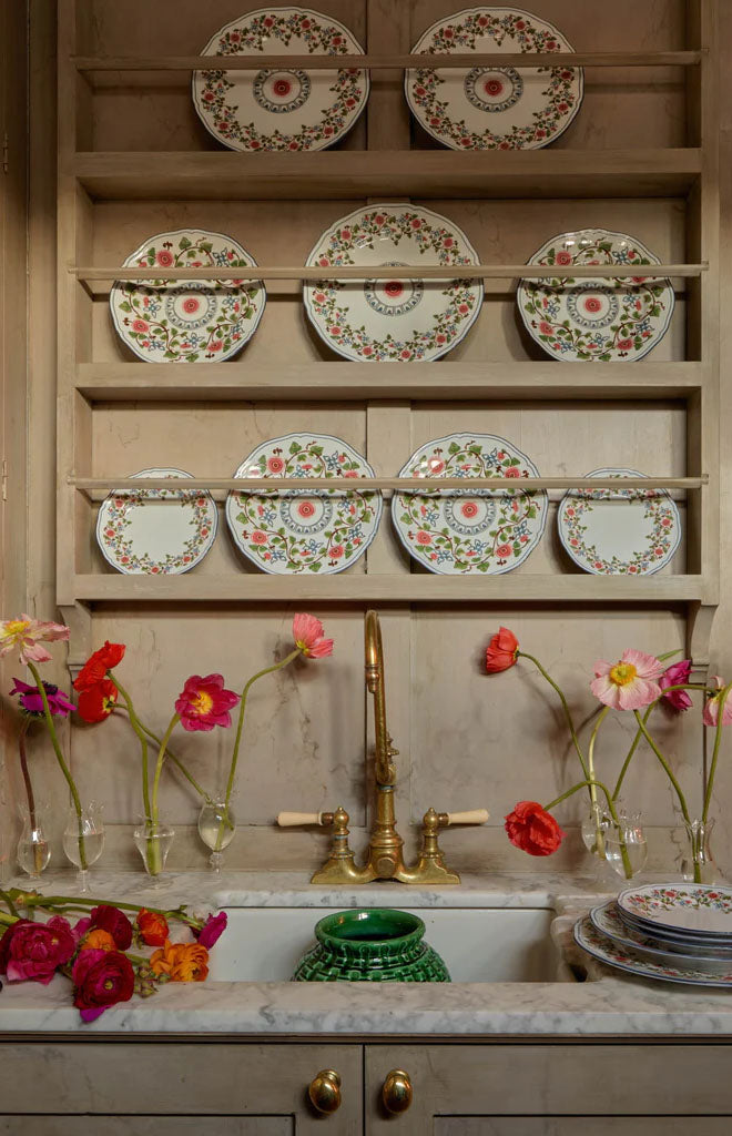 Ginori Martina charger plates arranged on a wooden plate rack above a marble sink with a brass faucet and pink poppy stems