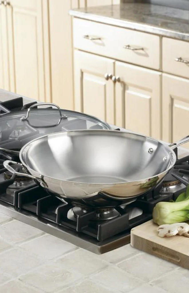 Three-quarter view of Chef’s Classic 14-inch stir-fry pan in polished stainless steel with helper handles and glass lid on stovetop
