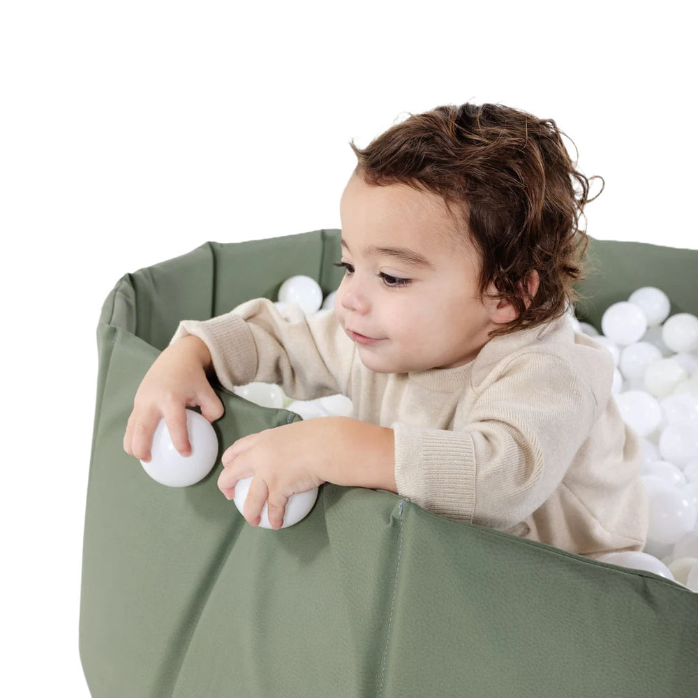 Three-quarter view of Children’s Ball Pit showcasing fabric walls filled with white play balls with a toddler leaning on the rim against a white background