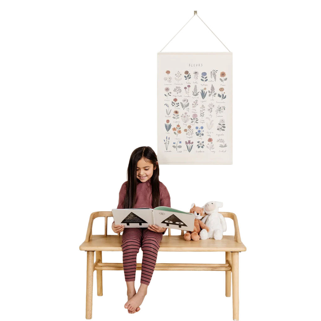 Girl sitting on a wooden bench reading a book with a floral Fleurs children’s poster hanging above and stuffed animals beside her
