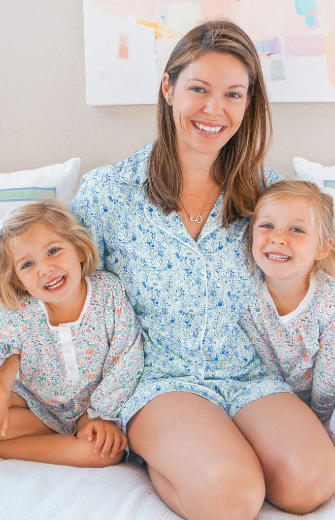Woman wearing blue floral short pajama set sitting on a bed with two smiling girls in coordinating prints