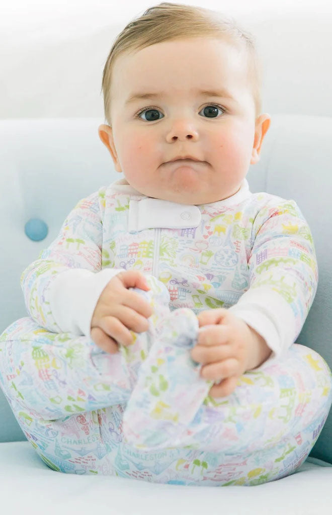 Front view of Charleston zip baby onesie on a seated baby showing full-zip front, footed legs, and pastel Rainbow Row print on a light chair