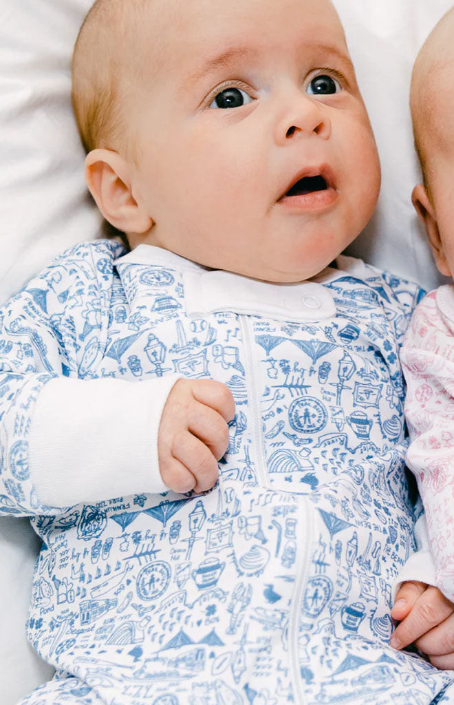Front view of Boston Zip Baby Onesie in Sailor Blue showcasing hand-drawn Beantown landmark print on a baby against white bedding