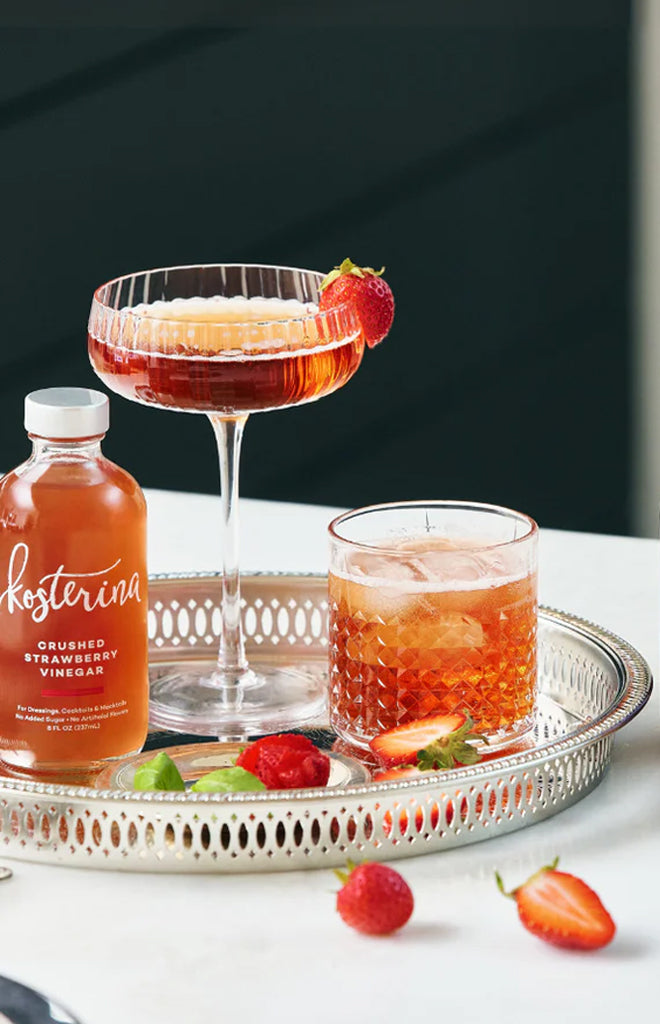 Front view of Kosterina crushed strawberry vinegar bottle with two cocktails and fresh strawberries on silver serving tray