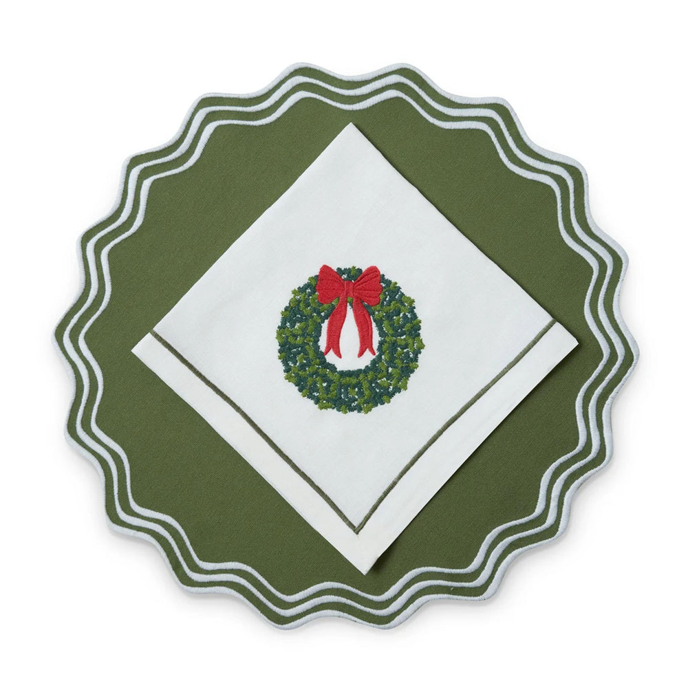 Top view of Wreath napkin folded on scalloped green charger showcasing embroidered wreath with red bow and stitched border
