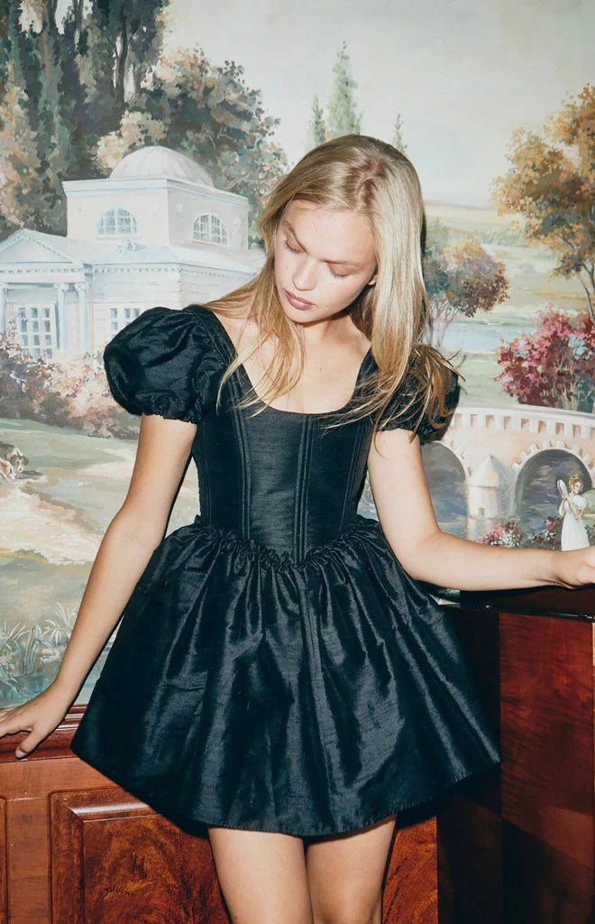 Front view of The Chloe dress in black dupioni on model: corseted bodice, puff sleeves, gathered mini skirt, mural backdrop