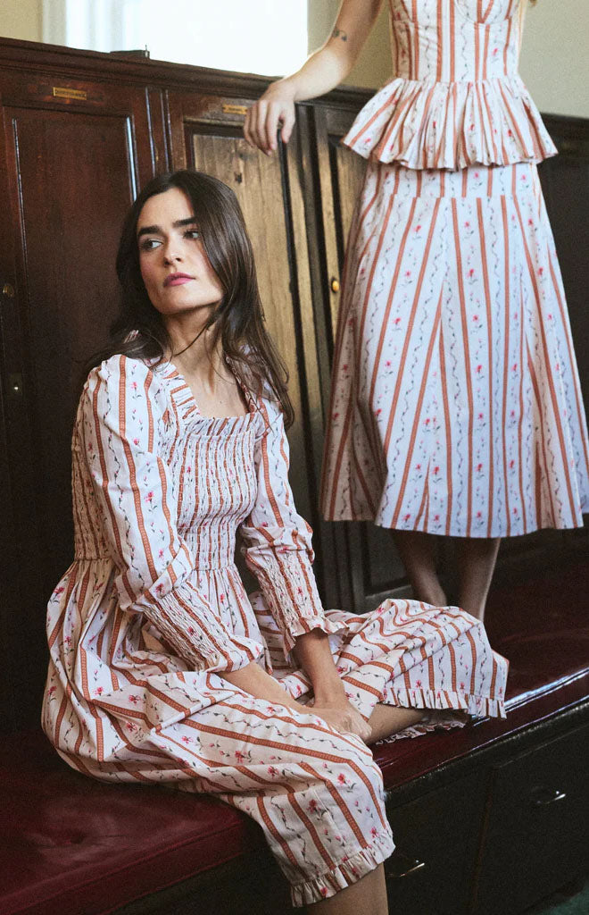 Front view of Eloise dress in Cherie showing smocked bodice, ruffled square collar and ankle ruffle on wooden bench