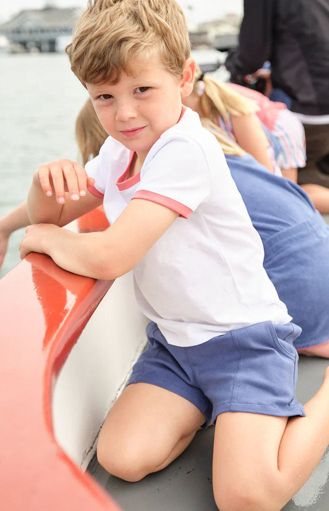 Boy kneeling on a boat wearing navy twill shorts and a white t-shirt with pink trim