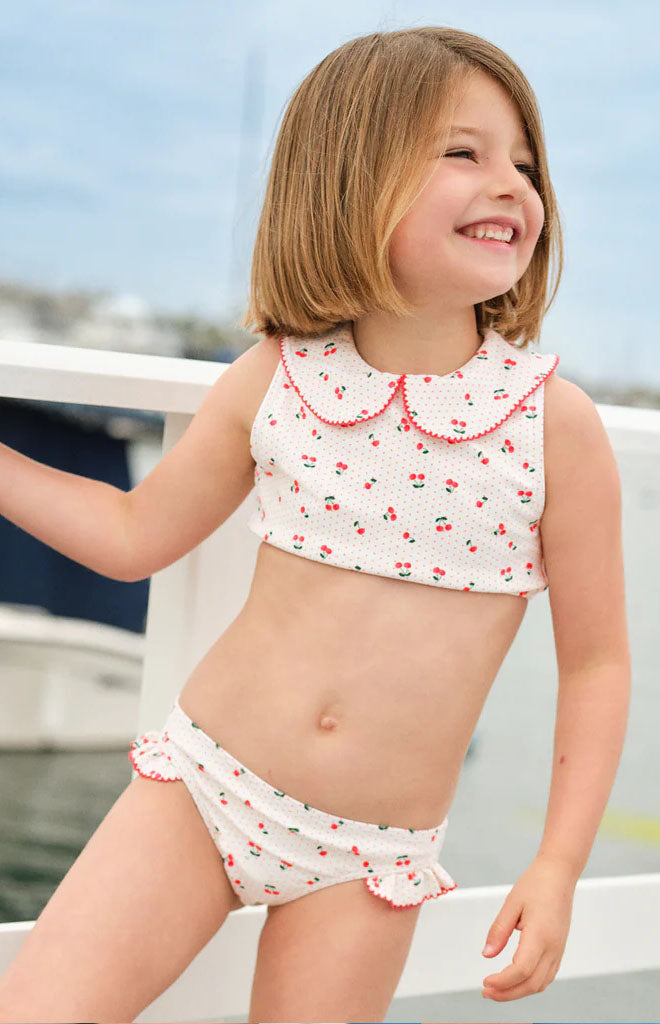 Front view of girls’ white cherry-print two-piece bikini with rounded scalloped collar and ruffle-trim bottoms, modeled at a marina