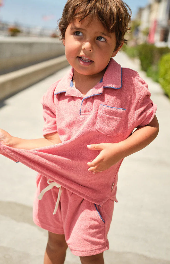 Young boy wearing poppy red French terry shorts and matching polo top pulling at his shirt on a sunny seaside promenade