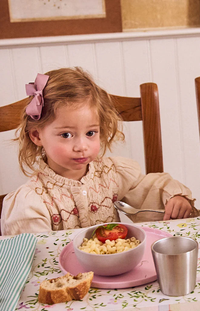 Young child sitting at a table eating from a gray bowl of pasta on a pink plate, holding a spoon