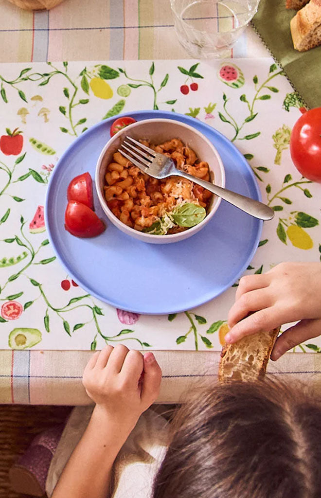 Child eating pasta at a table on Harvest Tangle placemat with blue plate, bowl of pasta, tomato wedges, and bread