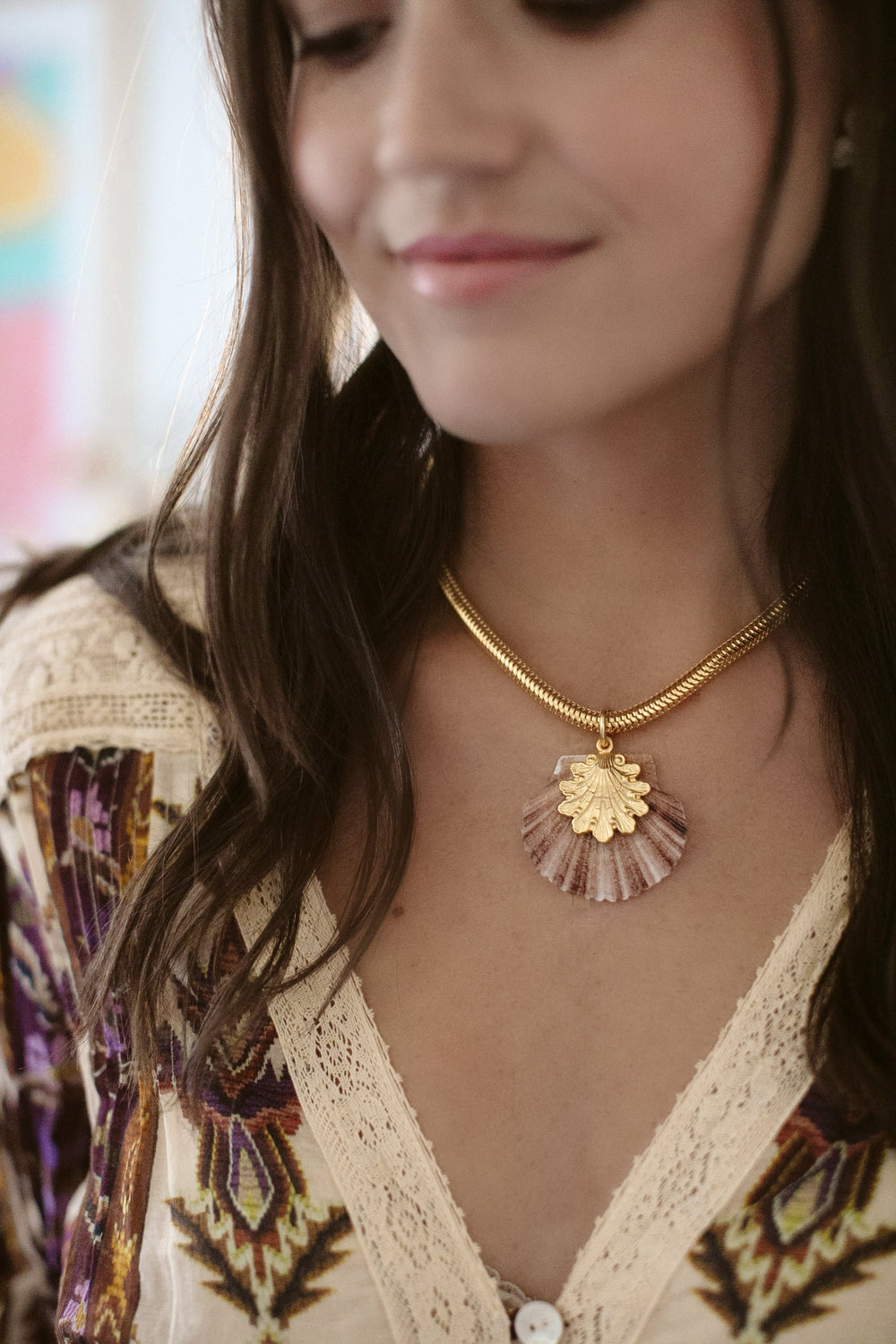Close-up of Oceana Necklace on model showcasing a baby scallop shell pendant with 24K antique gold plating on a gold snake chain, soft indoor background