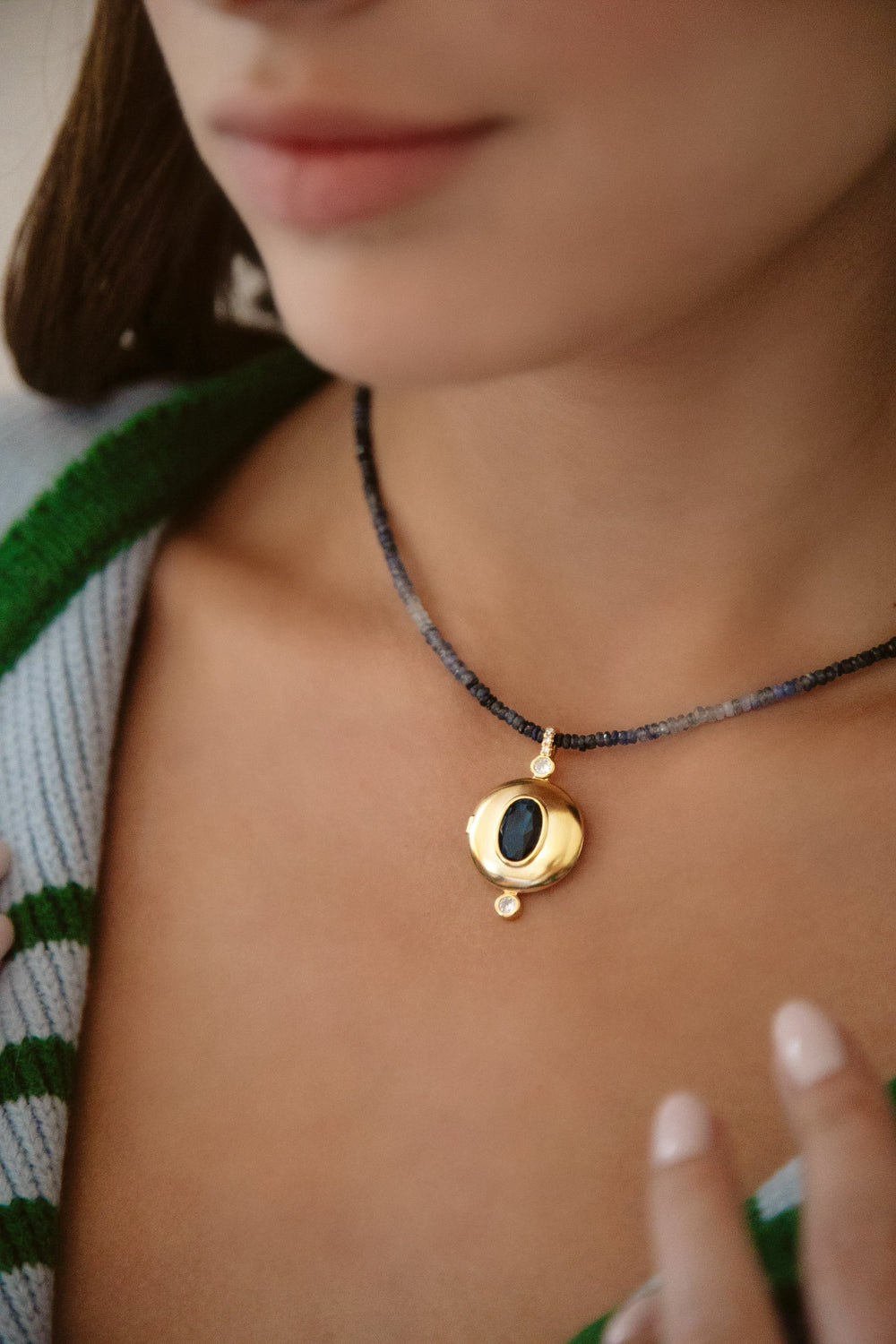 Close-up of Orion Necklace on model: faceted blue ombré sapphire beads with a 24K gold-plated round locket featuring a dark center stone