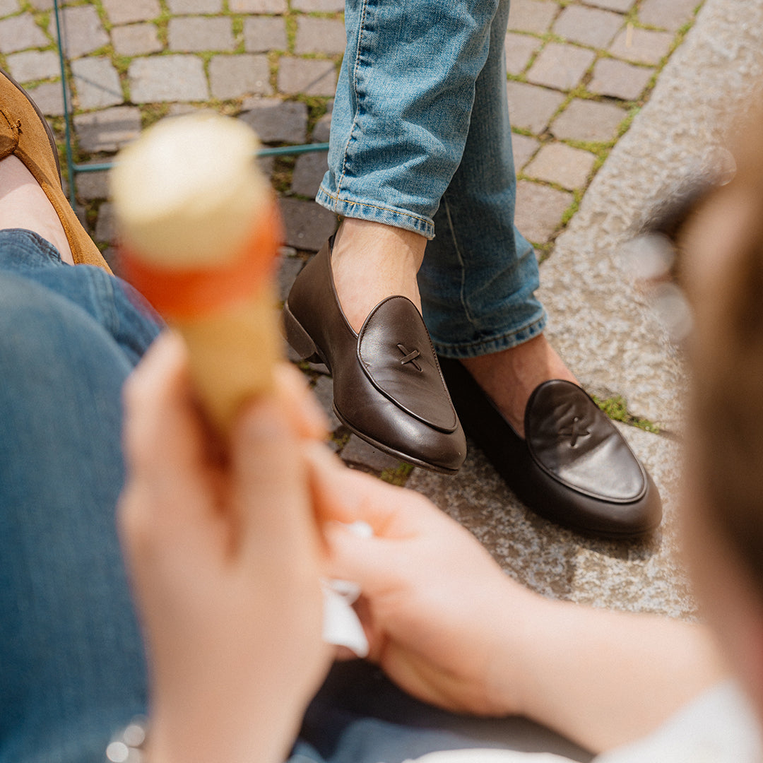 Man wearing brown Milano leather loafers sitting on a cobblestone curb with legs crossed, someone holding an ice cream cone in the foreground