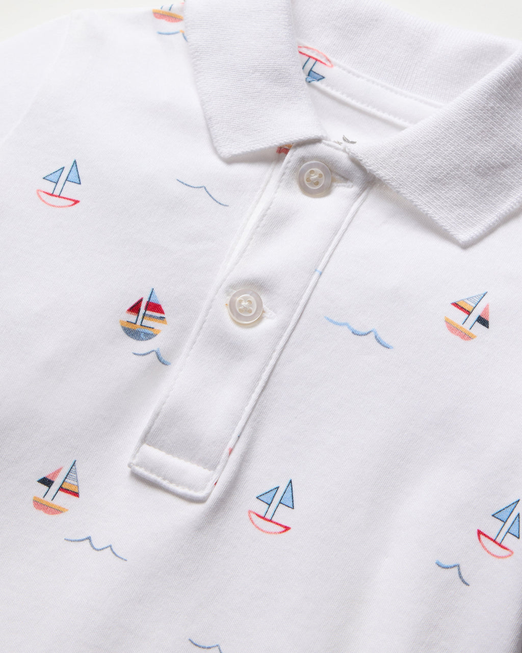 The Babys Pima Louis Play Romper in Bateau by Petite Plume is crafted from 100% Pima cotton, featuring a playful print with sailboats in blue, red, and yellow. It includes a classic collar and two-button placket, highlighted by wavy blue lines among the sailboat designs.