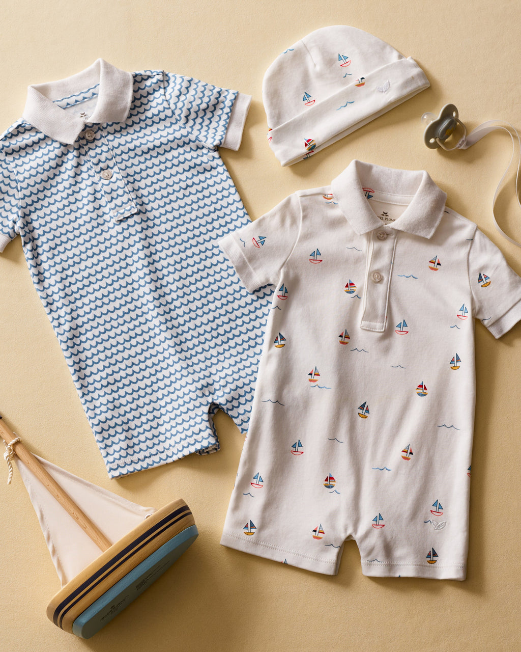 Displayed are two Petite Plume Pima cotton rompers: one with vibrant blue waves and a white collar, the other in a sailboat print. They are surrounded by a matching sailboat hat, pacifier, and wooden sailboat on a beige background.