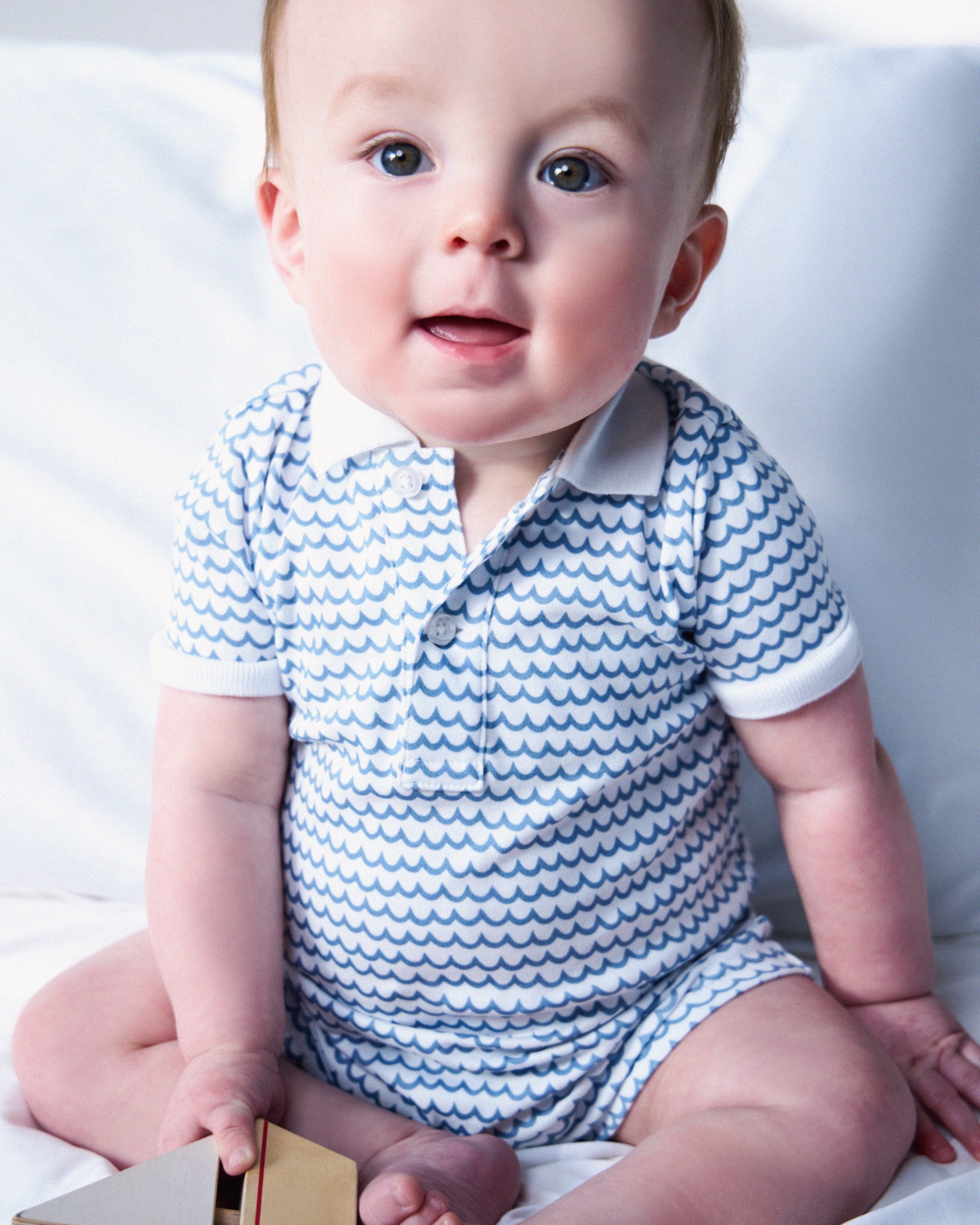 A baby in a white and blue patterned Petite Plume Babys Pima Louis Play Romper in La Mer sits on a white surface, holding a small object and gazing forward with an expression as lively as the sea.