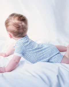 A baby with light brown hair lies on their stomach in serene comfort, wearing Petite Plumes Babys Pima Louis Play Romper in La Mer, featuring light blue wavy sea-inspired stripes, against a soft white background. The focus is on their back and head.